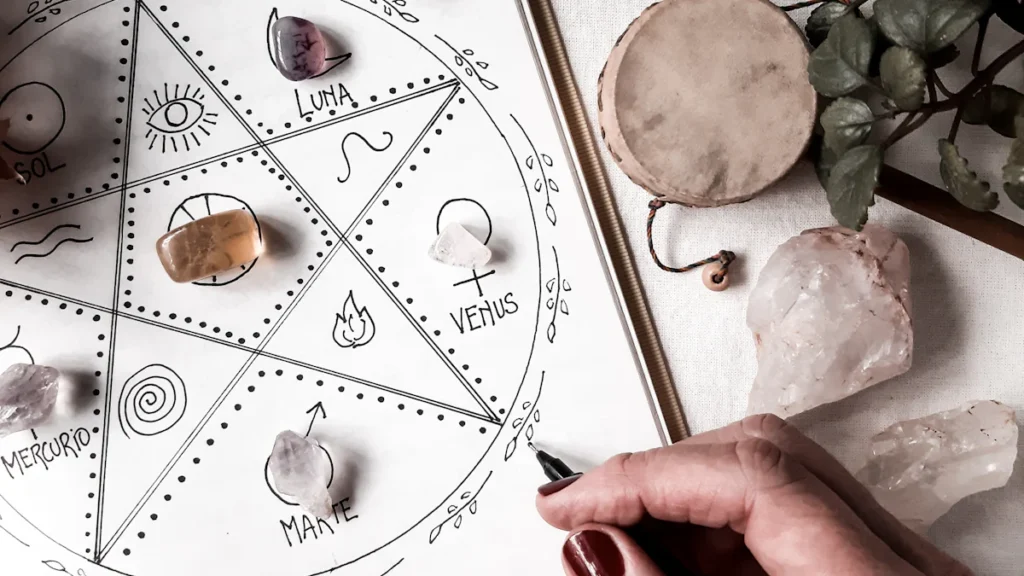 O mână ține un stilou peste o foaie împodobită cu simboluri astrologice, contemplând tranzitele astrologice și oportunitățile pe care le dezvăluie. Cristalele sunt așezate cu grijă în jur, în timp ce o mică tobă și o plantă aduc armonie acestui spațiu transformator.