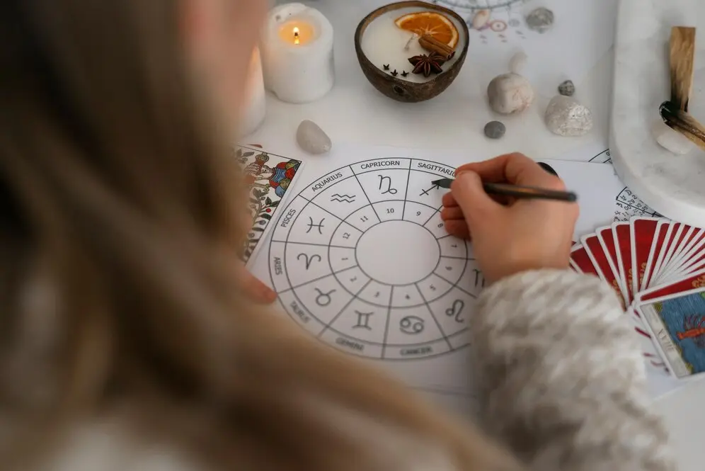 O persoană înscrie cu atenție note pe o diagramă zodiacală la o masă împodobită cu lumânări pâlpâitoare, pietre mistice și cărți de tarot, căutând să transforme tranzitele astrologice în oportunități de creștere.